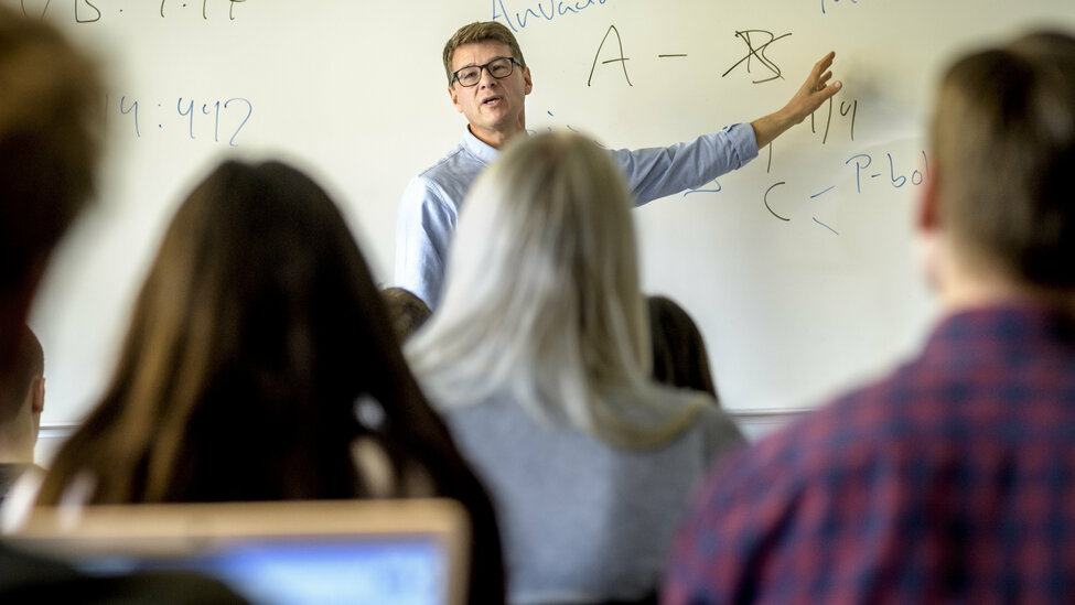 [Translate to English:] Lärare vid whiteboard i ett klassrum, framför studenter. 
