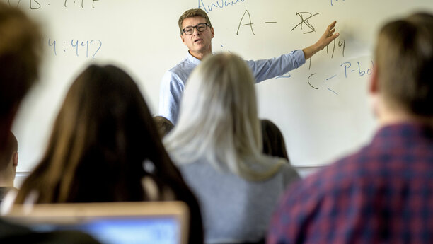Lärare vid whiteboard i ett klassrum, framför studenter. 